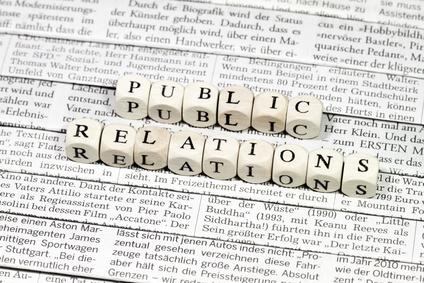 Die Zeichenkette "Public Relations" zusammengesetzt aus Buchstabenwürfeln vor gedrucktem Text